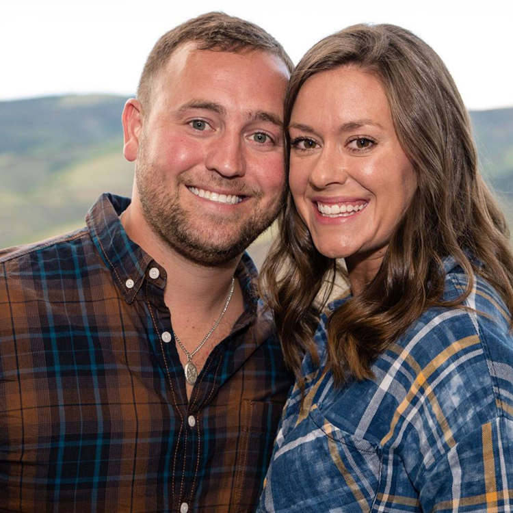 Two people smiling at camera wearing plaid shirts with scenic hillside background.