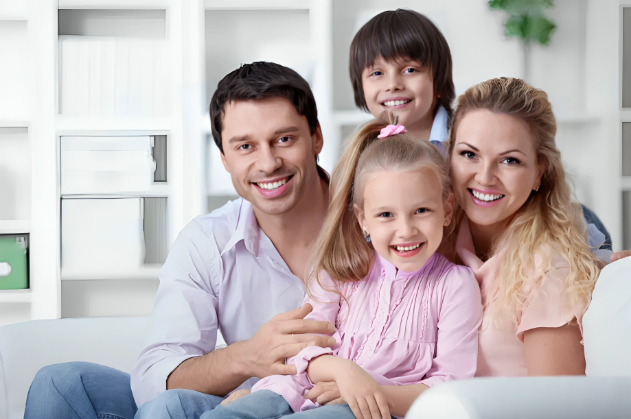 Happy family of four smiling together in modern home setting with white shelving background.