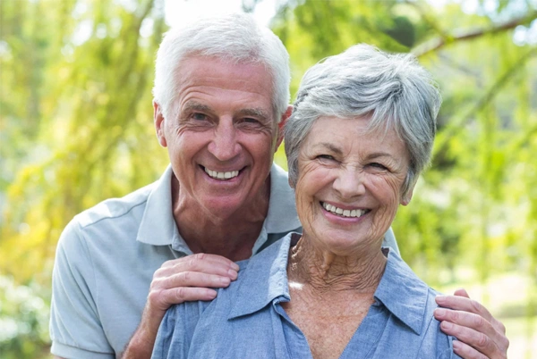 Smiling older couple outdoors in casual blue clothing with green foliage background.